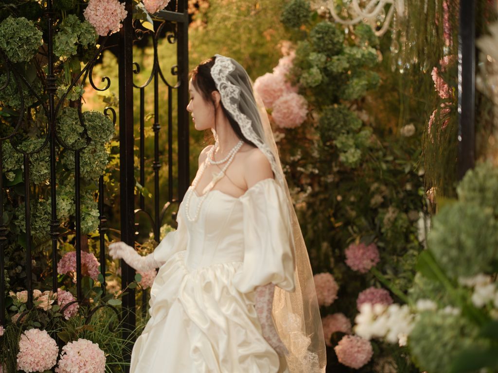 A bride in a vintage gown stands in a lush garden, surrounded by blooming hydrangeas.